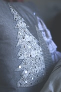 A close-up view of a textile surface featuring a triangular lace pattern, adorned with sequins. The lace is shaped like a stylized tree, with intricate detailing and reflective embellishments. The fabric is soft grey, providing a muted backdrop.