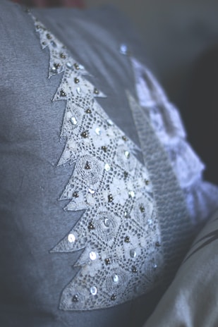 A close-up view of a textile surface featuring a triangular lace pattern, adorned with sequins. The lace is shaped like a stylized tree, with intricate detailing and reflective embellishments. The fabric is soft grey, providing a muted backdrop.