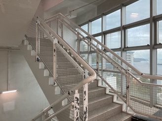 A stairway with metal railings and a grid-like pattern is seen inside a building. The walls and ceiling appear to be made of concrete, and several large windows line the right side, offering a view of an overcast sky and distant water. The paint on the railings is chipped, giving a slightly worn appearance.