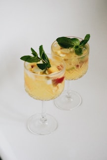 Two elegant glass goblets containing a light yellow beverage filled with ice cubes and garnished with fresh mint leaves and pieces of fruit. The glasses have a decorative, textured design and are placed against a smooth, white background.