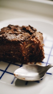 A close-up of a dense, chocolate dessert with a textured, slightly glossy surface, sitting on a white plate with blue grid lines, accompanied by a reflective metal spoon. The dessert appears rich and decadent, suggesting a homemade or artisanal quality.