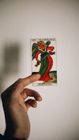 A hand holds a tarot card depicting an angelic figure with green and red robes. The card bears the title 'Temperance' with the number XIV at the top. The background is plain with a soft shadow cast by the hand and card.