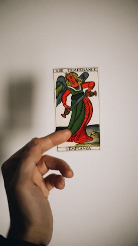 A hand holds a tarot card depicting an angelic figure with green and red robes. The card bears the title 'Temperance' with the number XIV at the top. The background is plain with a soft shadow cast by the hand and card.