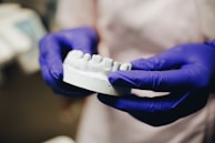 A pair of hands wearing purple gloves holds a white dental mold resembling a set of teeth. The background is blurred, focusing attention on the dental mold and the gloves.