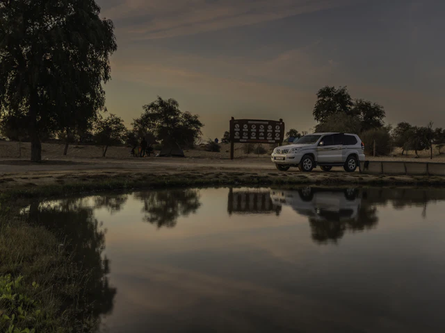 A 4x4 parked beside one of the Al Qudra desert lakes at dusk, its silhouette and the trees behind it mirrored in the still water.