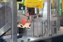 A section of an industrial machine, featuring metal components and a mechanism for processing or assembling parts. The setup includes wiring and a slanted metal panel, with a robotic element prominent in the background.