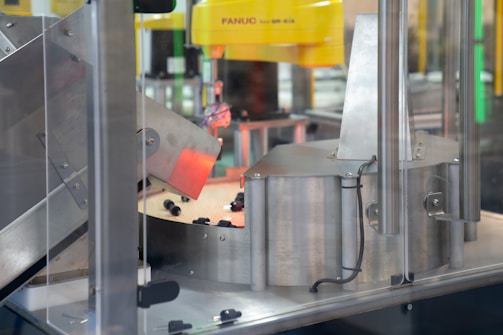 A section of an industrial machine, featuring metal components and a mechanism for processing or assembling parts. The setup includes wiring and a slanted metal panel, with a robotic element prominent in the background.
