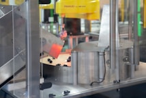 A section of an industrial machine, featuring metal components and a mechanism for processing or assembling parts. The setup includes wiring and a slanted metal panel, with a robotic element prominent in the background.