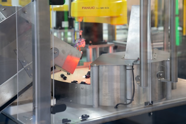A section of an industrial machine, featuring metal components and a mechanism for processing or assembling parts. The setup includes wiring and a slanted metal panel, with a robotic element prominent in the background.
