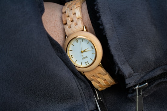 A close-up of a wooden wristwatch on a person's wrist. The watch has a round face with a light-colored dial and black numbers. The wristband is made of interlocking wooden links, and the person is wearing a dark sleeve.