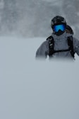 A person wearing winter sports gear, including a helmet and goggles, stands in a snowy landscape. The snow is falling heavily, partially obscuring the lower part of the body. The environment appears cold and overcast.