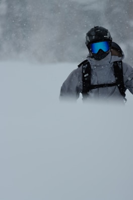 A person wearing winter sports gear, including a helmet and goggles, stands in a snowy landscape. The snow is falling heavily, partially obscuring the lower part of the body. The environment appears cold and overcast.