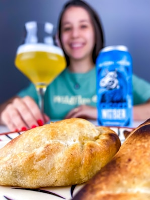 A close-up view of golden-brown pastries on a plate, with a smiling person in the background holding a glass of beer and a blue beverage can. The focus is on the food in the foreground.