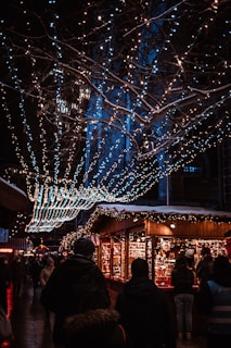A bustling nighttime market scene features wooden stalls decorated with twinkling string lights. Several people are browsing and walking around, bundled up in winter clothing. The atmosphere is festive, with intricate light displays hanging from trees overhead, creating an enchanting ambiance.