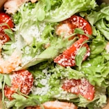A close-up view of a fresh salad with leafy greens, sliced red tomatoes, and grated white cheese sprinkled on top. Some shrimp pieces are visible, adding protein to the dish.