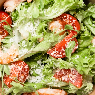 A close-up view of a fresh salad with leafy greens, sliced red tomatoes, and grated white cheese sprinkled on top. Some shrimp pieces are visible, adding protein to the dish.