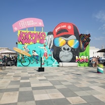 A vibrant mural covers the wall featuring a large gorilla wearing sunglasses and a red cap with writing on it. The mural includes graffiti elements such as the word 'Welcome' and showcases figures participating in sports like cycling and running. Bright colors and artistic elements, including a surfboard and paint roller, contribute to the dynamic scene. The area in front of the mural consists of a tiled plaza with some people visible.