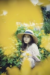 A sunlit scene of a woman in a flowing floral dress and wide-brimmed hat standing in a blooming garden.