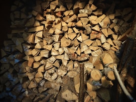 A neatly stacked pile of chopped firewood, with logs cut into uniform, triangular wedges. The logs have a natural, beige color with some darker bark still attached.