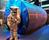 An astronaut suit displayed in front of a large, cylindrical structure with a metallic surface and geometric patterns. The setting appears to be an exhibition or display area with vibrant lighting.