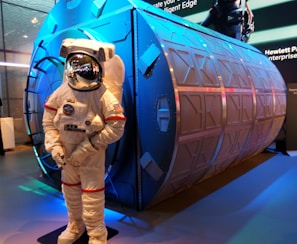 An astronaut suit displayed in front of a large, cylindrical structure with a metallic surface and geometric patterns. The setting appears to be an exhibition or display area with vibrant lighting.