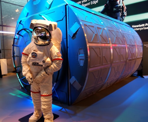 An astronaut suit displayed in front of a large, cylindrical structure with a metallic surface and geometric patterns. The setting appears to be an exhibition or display area with vibrant lighting.