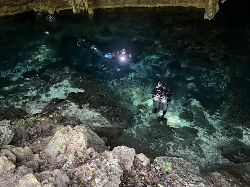 Two scuba divers are exploring a clear, shallow body of water inside a cave. The divers are equipped with diving gear and carry underwater lights, which illuminate their path. The rocky cave ceiling and walls are visible above the water, and the ground is covered in rocks and pebbles.