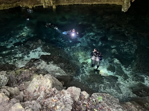 Two scuba divers are exploring a clear, shallow body of water inside a cave. The divers are equipped with diving gear and carry underwater lights, which illuminate their path. The rocky cave ceiling and walls are visible above the water, and the ground is covered in rocks and pebbles.