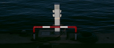 A waterside scene features a metal post beside the water, surrounded by a red and white striped barrier. A coil of cable lies next to the barrier, resting on the concrete surface near the edge of the water.