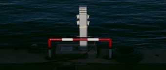 A waterside scene features a metal post beside the water, surrounded by a red and white striped barrier. A coil of cable lies next to the barrier, resting on the concrete surface near the edge of the water.