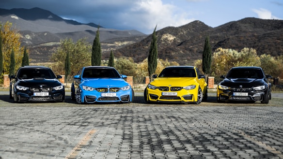 Four sports cars are parked in a row on a paved area with a backdrop of mountains and tall cypress trees. The cars are in different colors, including yellow, blue, and two black, and they have sleek, modern designs. The scene is set outdoors with a partly cloudy sky.