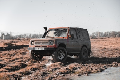 A rugged off-road vehicle drives through a muddy and grassy terrain. The car is covered in dirt, and water splashes around as it moves. The background features an open field with sparse trees and a clear sky.