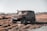 A rugged off-road vehicle drives through a muddy and grassy terrain. The car is covered in dirt, and water splashes around as it moves. The background features an open field with sparse trees and a clear sky.