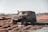 A rugged off-road vehicle drives through a muddy and grassy terrain. The car is covered in dirt, and water splashes around as it moves. The background features an open field with sparse trees and a clear sky.