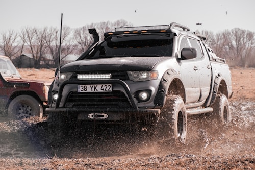 A rugged, black pickup truck navigating through a muddy terrain with splashes of water and mud flying around. The truck has modifications for off-road purposes, such as large tires and additional lights. In the background, another vehicle is partially visible, and there are leafless trees and an open field.