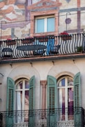 A building facade features artistic frescoes and a balcony adorned with black wrought-iron railings. Red flowers add a vibrant touch to the railing. Below, there are two arched windows with green shutters flanking a decorative column.