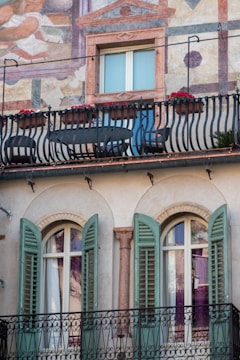A building facade features artistic frescoes and a balcony adorned with black wrought-iron railings. Red flowers add a vibrant touch to the railing. Below, there are two arched windows with green shutters flanking a decorative column.