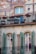 A building facade features artistic frescoes and a balcony adorned with black wrought-iron railings. Red flowers add a vibrant touch to the railing. Below, there are two arched windows with green shutters flanking a decorative column.
