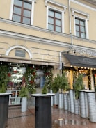 A street view of a building with a cream-colored facade, featuring large windows and decorative green foliage around the entrance. The canopy of a café or a shop is visible, displaying signage with the word 'Croissants'. Decorative lights and festive greenery adorn the exterior, suggesting a holiday theme.
