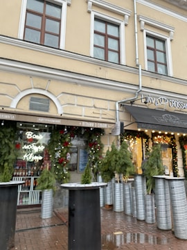 A street view of a building with a cream-colored facade, featuring large windows and decorative green foliage around the entrance. The canopy of a café or a shop is visible, displaying signage with the word 'Croissants'. Decorative lights and festive greenery adorn the exterior, suggesting a holiday theme.