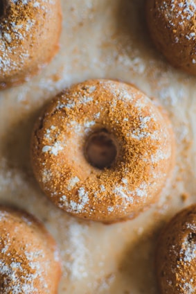 Several baked donuts are arranged on a surface, sprinkled with a light dusting of powdered sugar. The donuts have a golden brown color and appear freshly baked, with a soft texture visible on their surface.