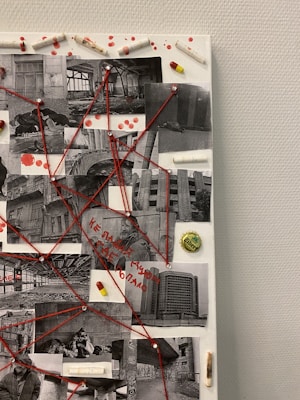 A collage of black-and-white photographs connected by red strings is displayed on a textured white board. The photographs depict urban decay, such as abandoned buildings and derelict interiors. Red push pins secure the strings to the board, forming a complex network. Scattered around the photographs are pills and capsules, as well as drops of red paint suggesting blood. In the center, Cyrillic text is visible, although its exact meaning is not clear.