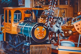 A colorful amusement park ride featuring a small, cheerful train engine with a smiling face on its front. Next to the train is a carousel horse painted in a glossy white finish. Bright and vibrant background elements include parts of other park rides, such as a small car and candy-striped decor.