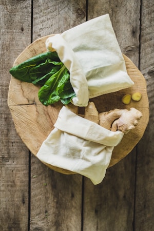 A wooden surface features a rustic cutting board with two cloth bags, one filled with fresh green leaves and the other with pieces of ginger. The wooden texture adds a natural, earthy feel to the composition.