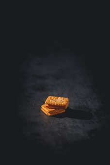 Two golden rectangular biscuits are stacked on a dark, textured surface. The biscuits feature intricate patterns on their top surfaces, casting soft shadows on the backdrop.
