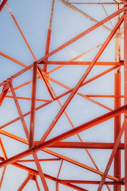 A complex network of orange steel beams intersecting at various angles against a clear blue sky, demonstrating an intricate structural framework.
