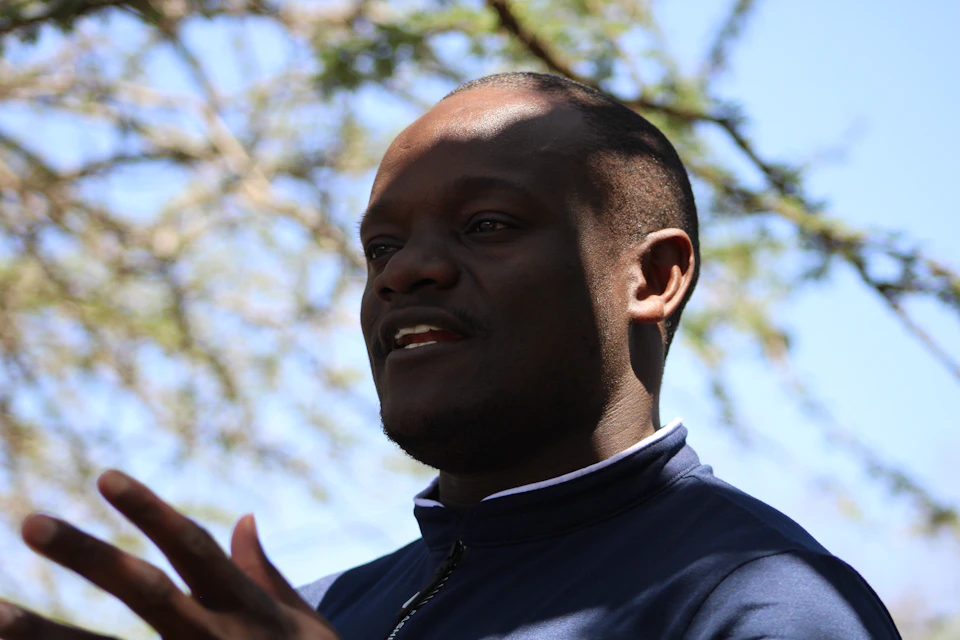 The image shows a man with short hair and a beard wearing a blue jacket, standing under a tree against a clear sky. The man is looking directly at the camera, engaging the viewer with his gaze.