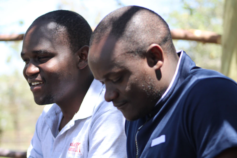 The image shows two men sitting next to each other on a bench, both looking off to the side with their eyes closed and mouths slightly open. The man on the left is wearing a white shirt with red text, while his companion on the right has a blue shirt. They are positioned in front of a tree, which adds a natural element to the scene.