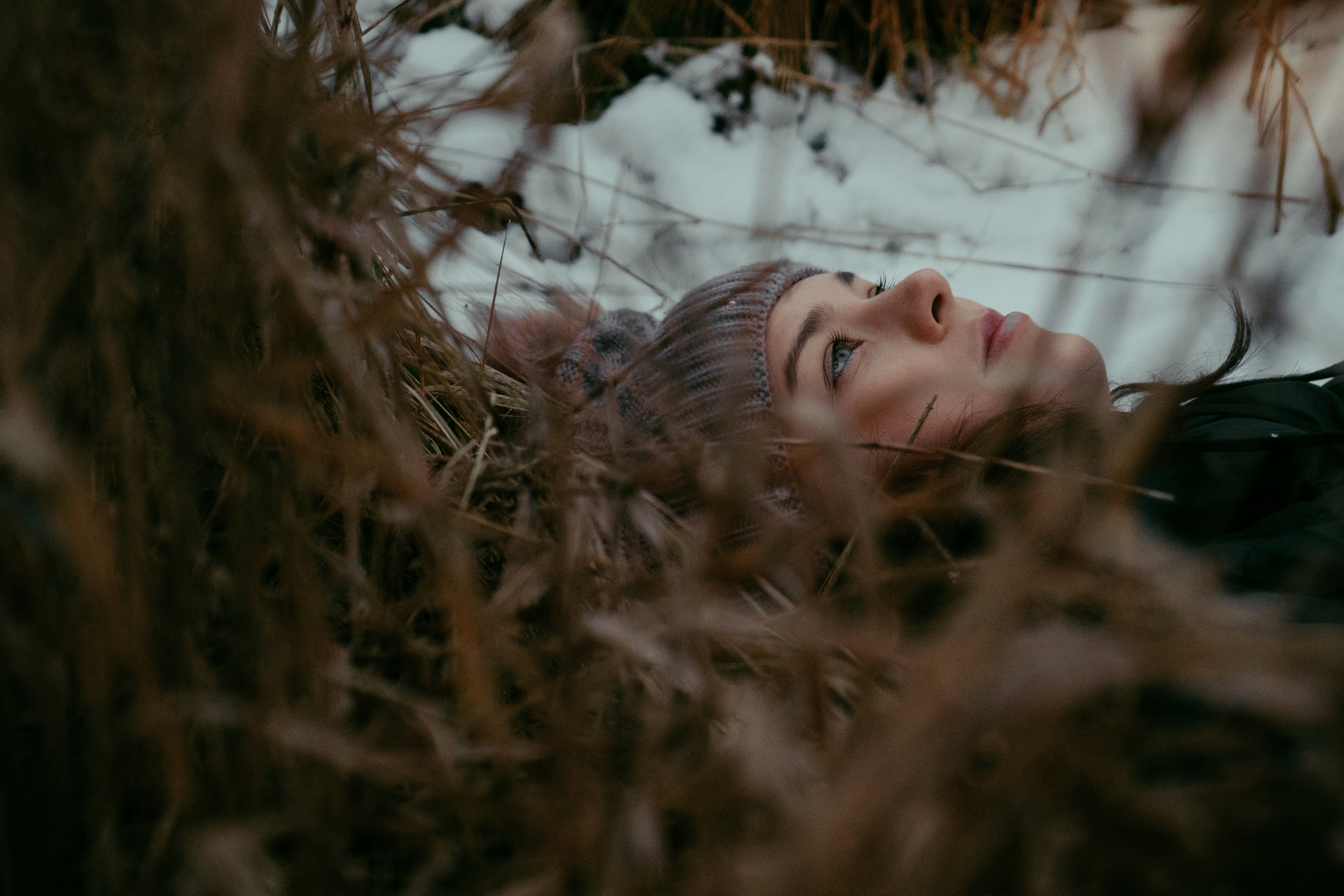 A white woman wearing a gray hat lies among long brown grass, snow in the background