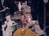 A collection of statues and sculptures in various styles, including figures of deities and historical figures. There is a prominently placed colorful poster, surrounded by stone carvings in a cluttered arrangement. The setting suggests an antique or collectible shop, with a mix of classical and cultural artifacts.